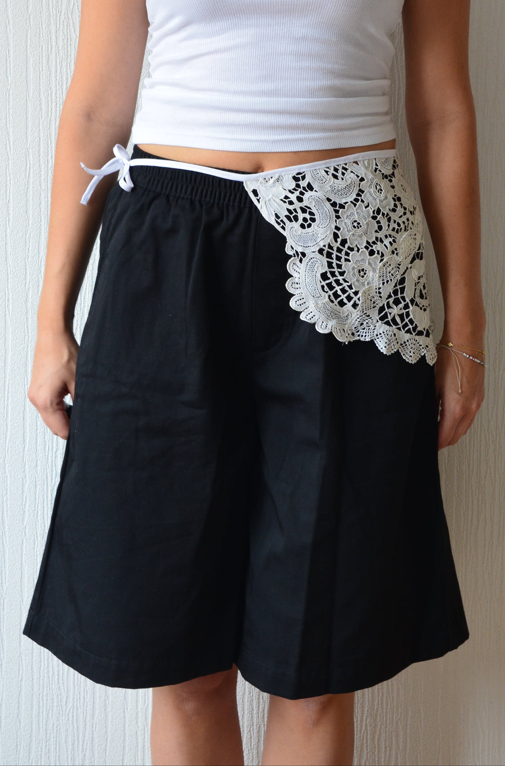 A person wearing black wide-leg shorts with a white lace panel and lace belt.