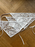 White lace bunting scarfs with white ties, laid out on a wooden floor. Two tags are attached to the center.