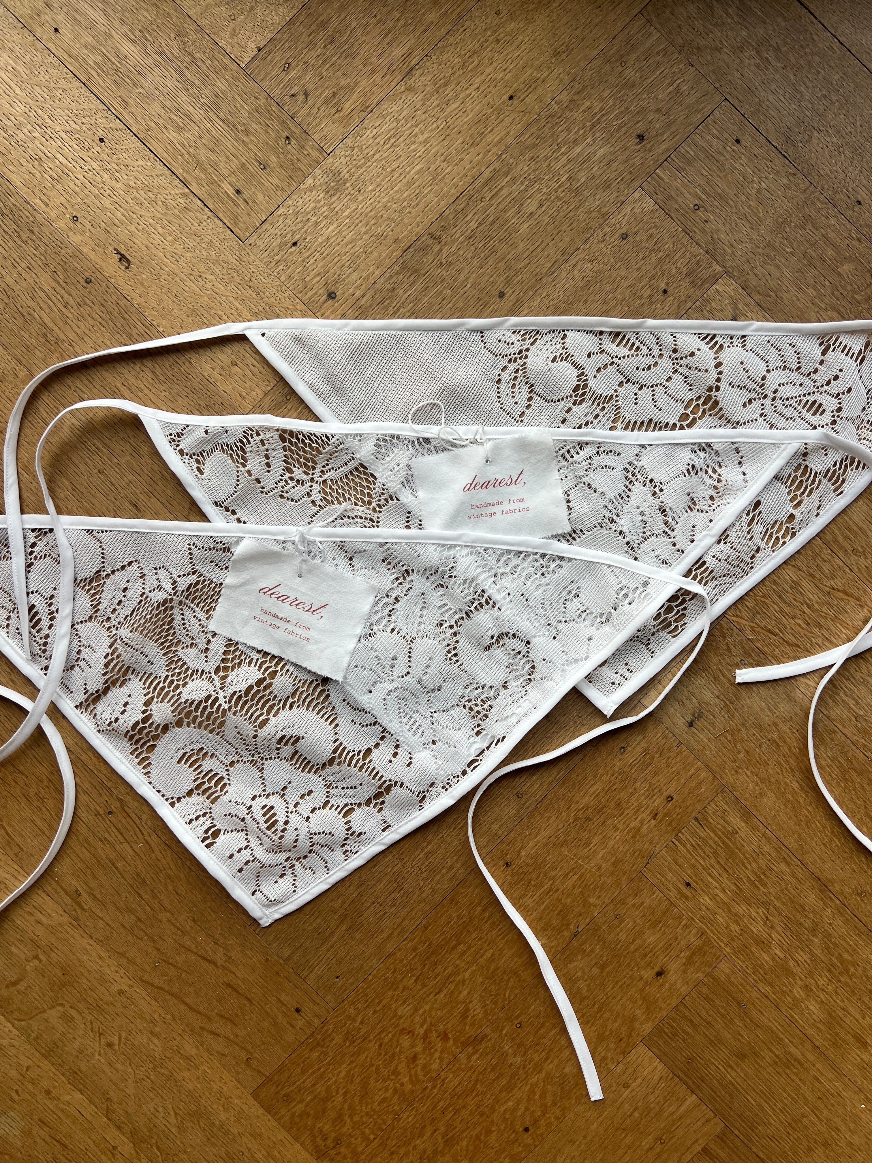 White lace bunting scarfs with white ties, laid out on a wooden floor. Two tags are attached to the center.