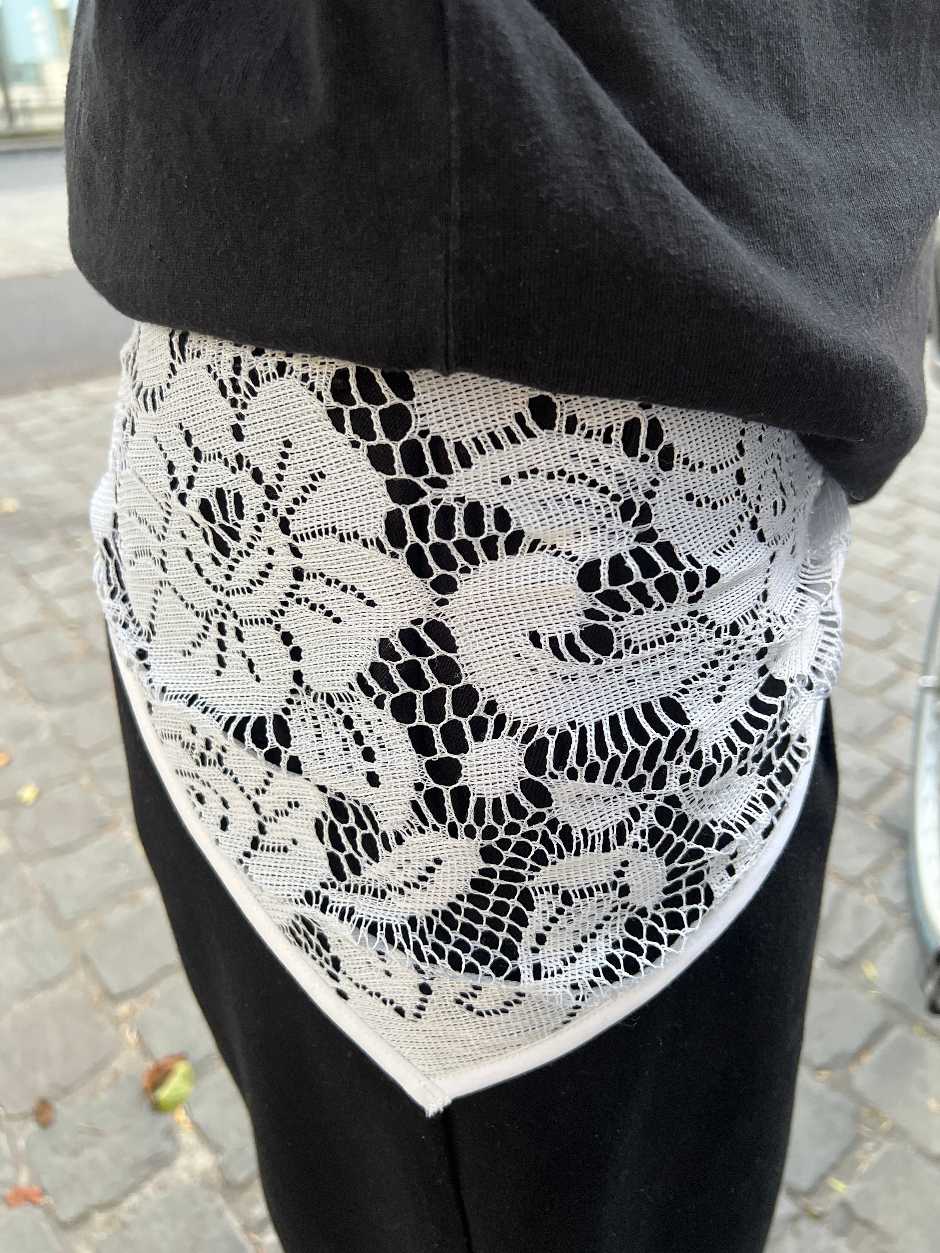 A close-up of a black garment showing a white floral lace overlay or cuff detail, set against an outdoor paved background.