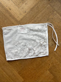 A white fabric face mask with an intricate floral lace pattern, laid flat on a wooden floor. It features a drawstring closure at the top and a small white brand tag near the bottom left.