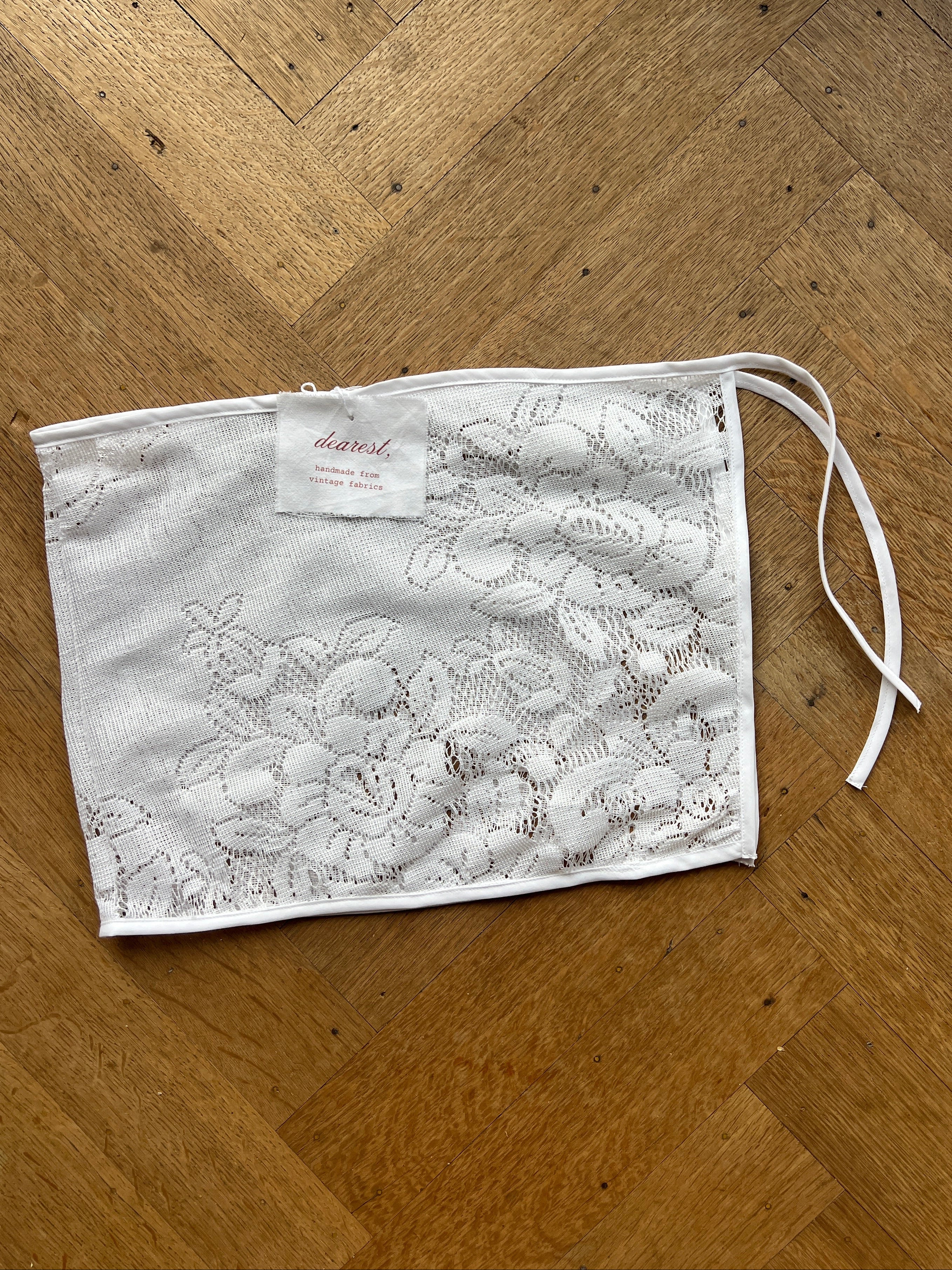 A white fabric face mask with an intricate floral lace pattern, laid flat on a wooden floor. It features a drawstring closure at the top and a small white brand tag near the bottom left.