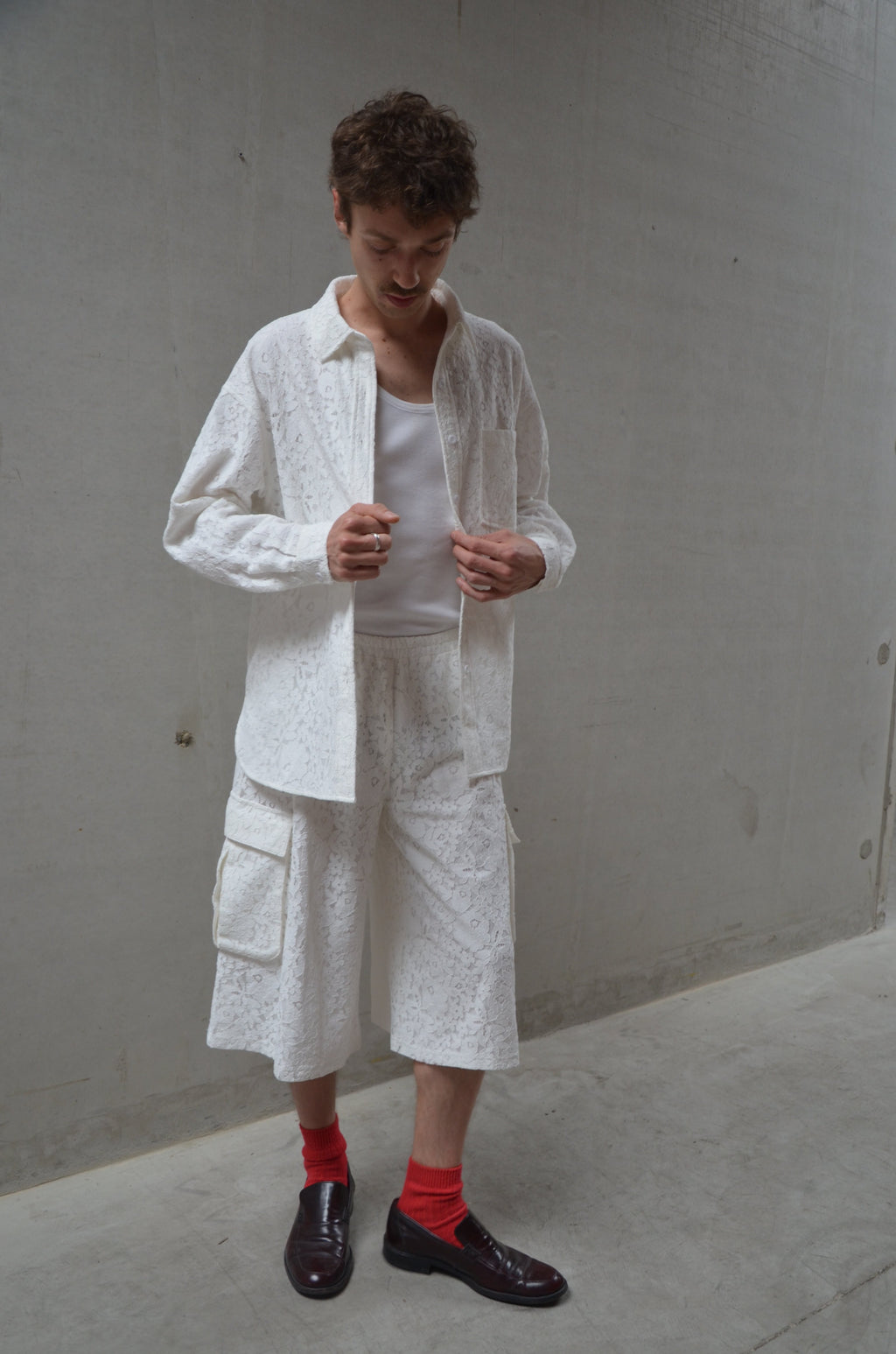 Person wearing a white lace shirt and shorts with red socks, standing against a plain wall.