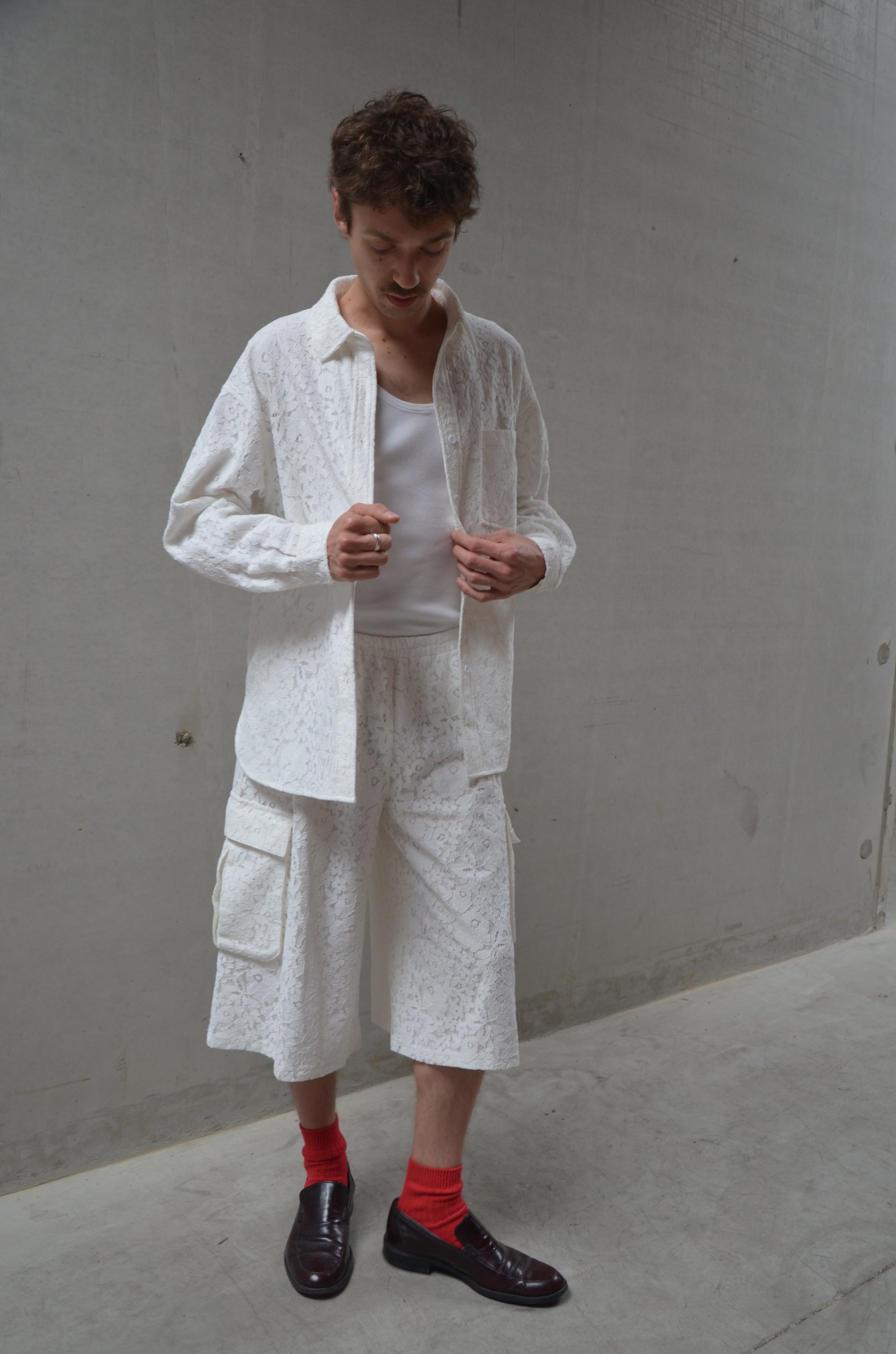 Person wearing a white lace shirt and shorts with red socks, standing against a plain wall.