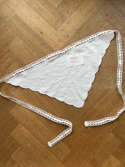 A white triangular bandana with scalloped edges and a long adjustable tie, laid flat on a wooden parquet floor. A small paper brand tag is attached near the center.